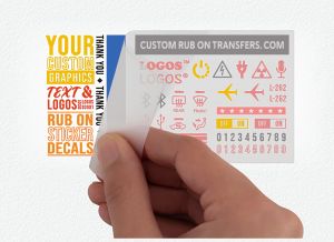 A hand peeling away a transparent transfer sheet from a white background. The Multi-Color Ensemble™ Transfer sheet ready to transfer in 1 application contains various red, yellow, and black custom graphics, texts, and logos including an airplane, audio waves, and numbers. The text "CUSTOM RUB ON TRANSFERS.COM" graces the top of this ready-to-transfer sheet.